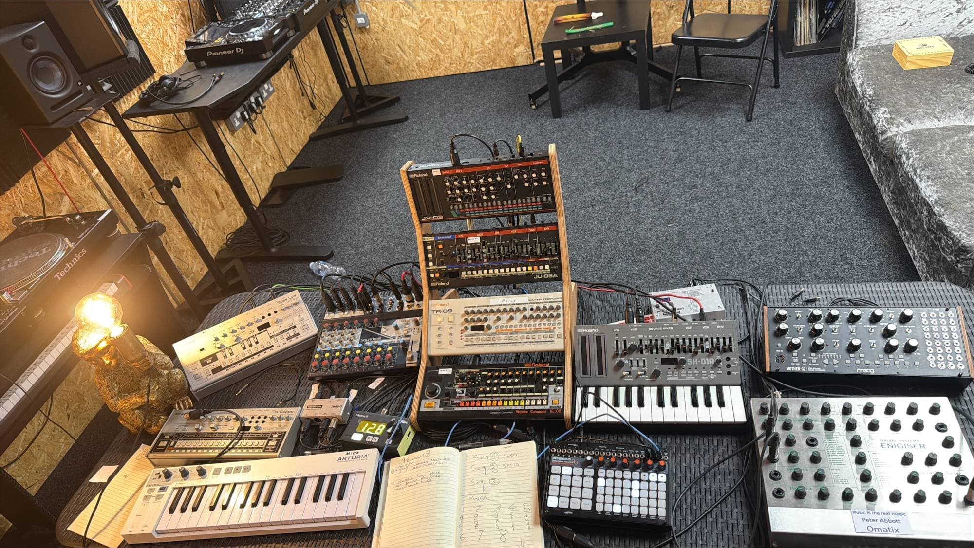 The synth table from above: a Roland Boutique rack, SH-01A, Moog Mother-32 and drum machines, with the DJ booth behind